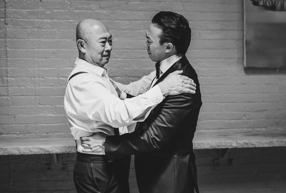 Two men share an emotional embrace against a brick wall in a black and white photo.