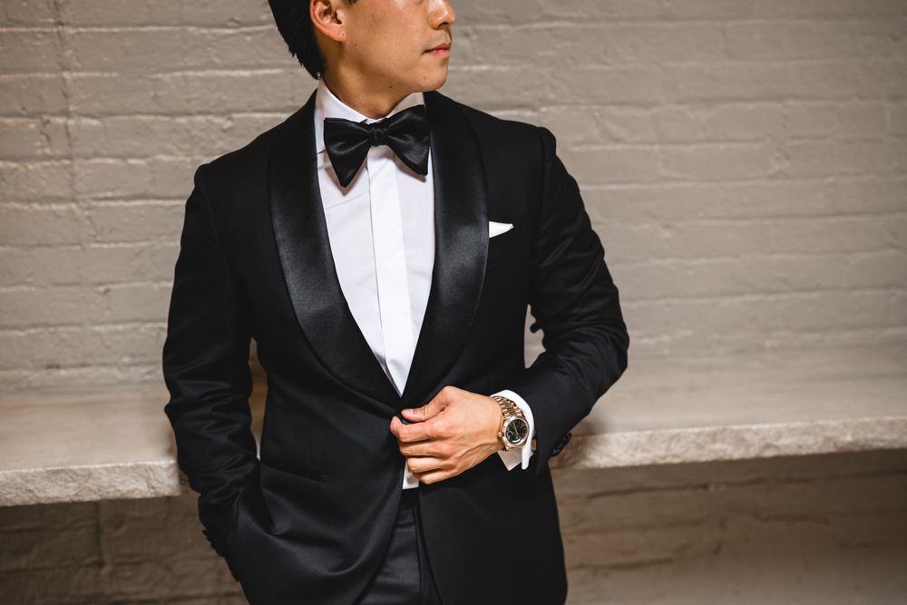 Groom in black tuxedo with bow tie and white pocket square posing against brick wall.