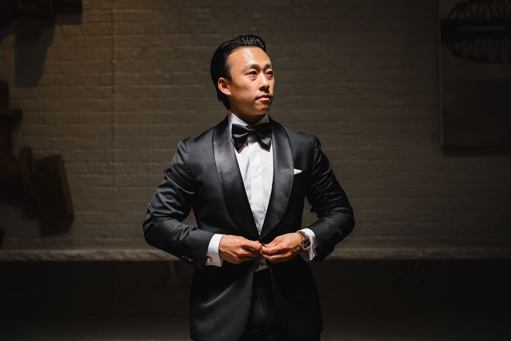Asian man in black tuxedo with bow tie buttoning jacket in dimly lit room.