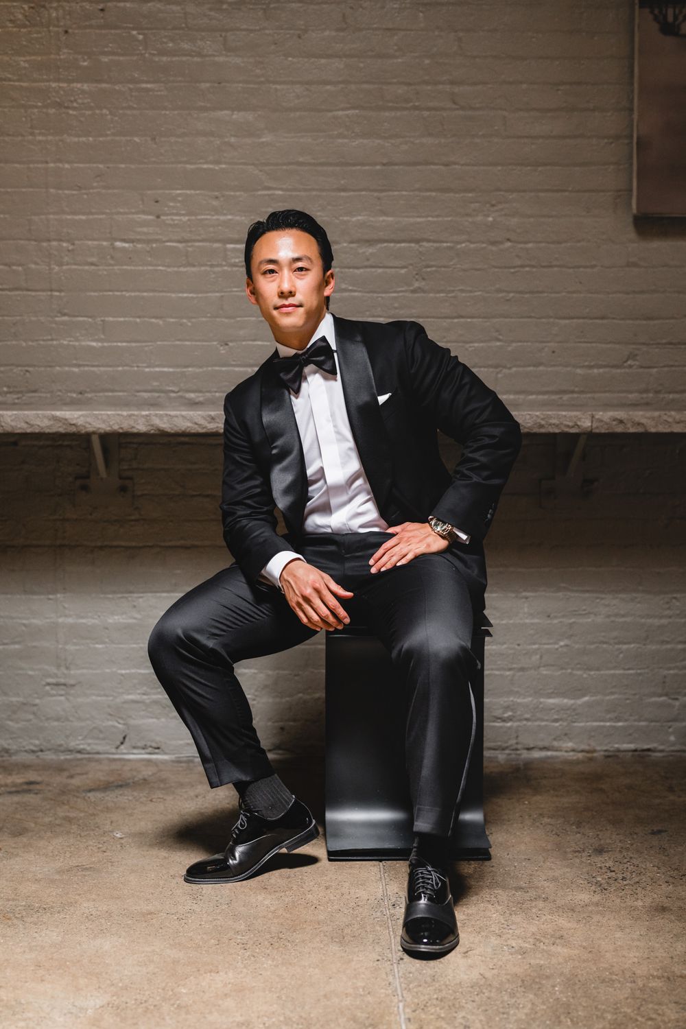 A man in a black tuxedo with bow tie sits confidently on a box against a white brick wall.