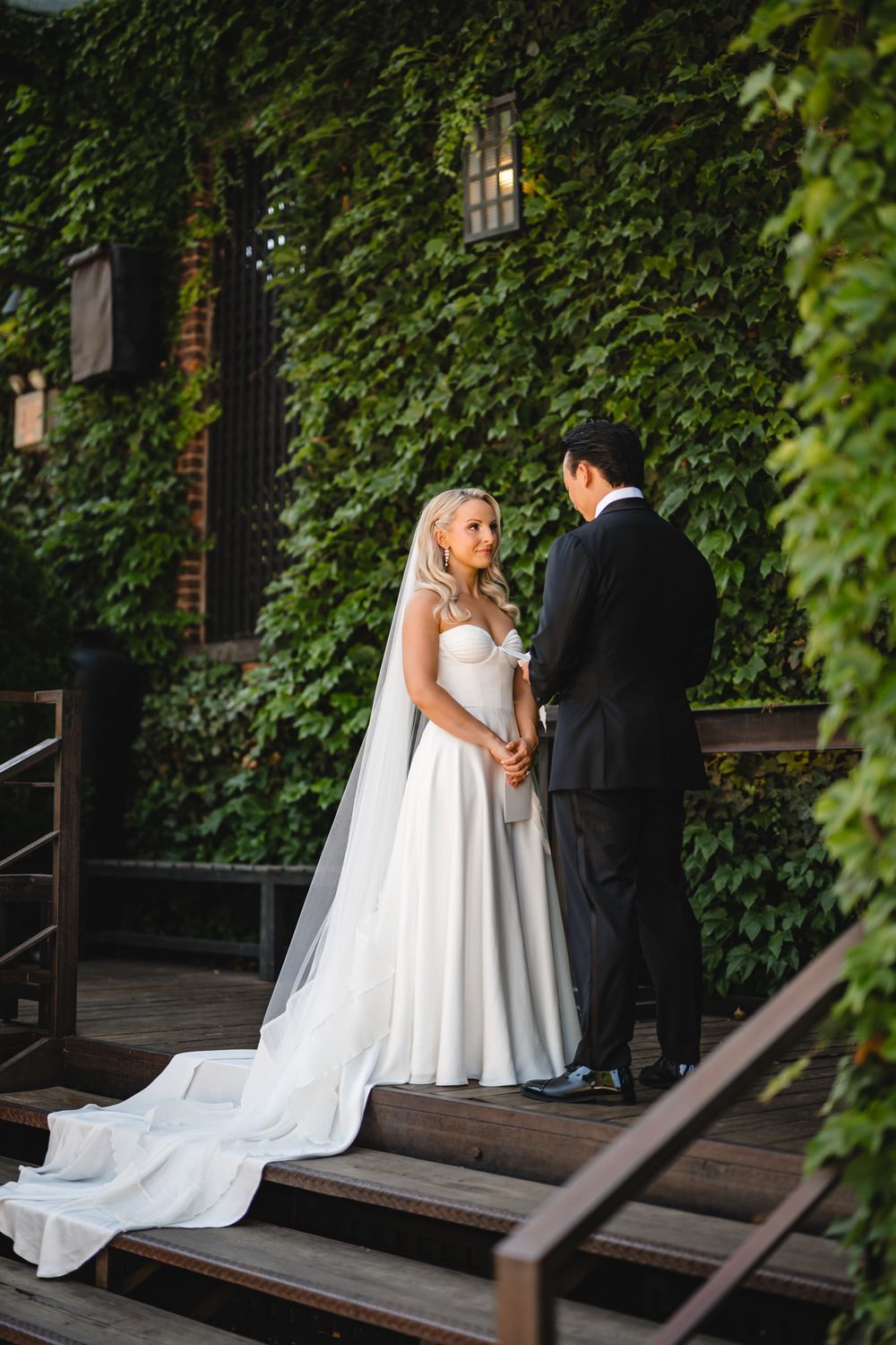 Bride and groom share vows on wooden steps surrounded by lush ivy-covered walls during outdoor wedding ceremony.