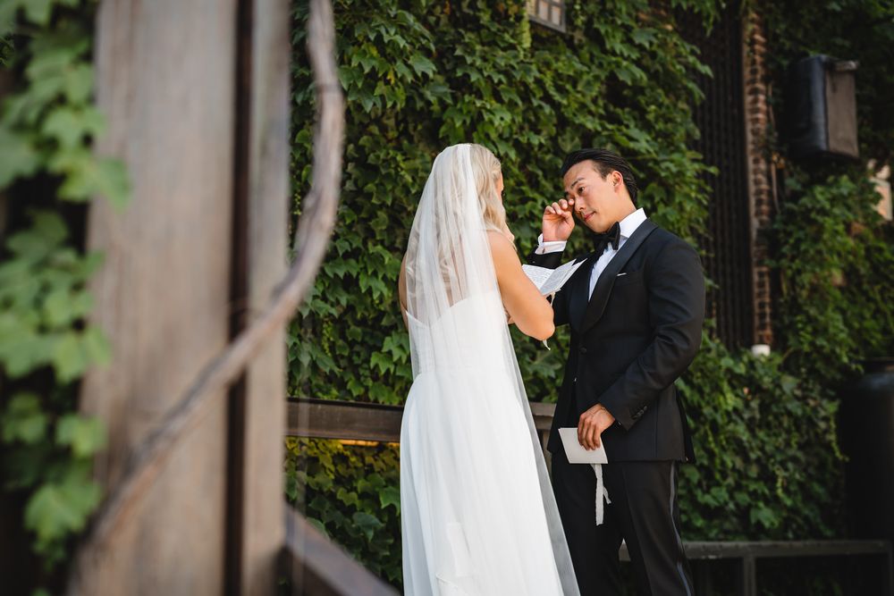 Groom in tuxedo wiping tears during emotional first look with bride in white gown and veil outdoors.