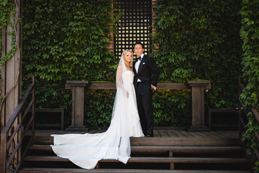 Bride and groom pose elegantly on wooden steps surrounded by lush ivy walls in a romantic outdoor wedding portrait.