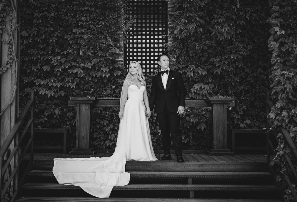 Elegant black and white wedding portrait of bride and groom standing on steps surrounded by lush ivy walls.