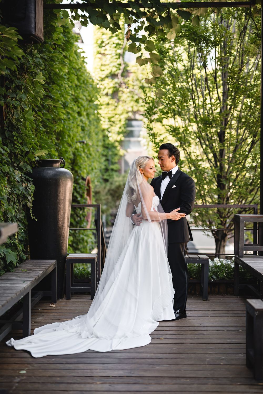 Newlyweds share an intimate moment on a sunlit wooden terrace surrounded by lush green foliage on their wedding day.