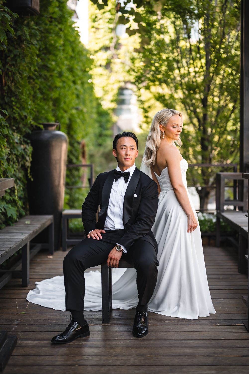 A groom in a black tuxedo sits on a chair while his bride in a white gown stands behind him on a wooden deck.