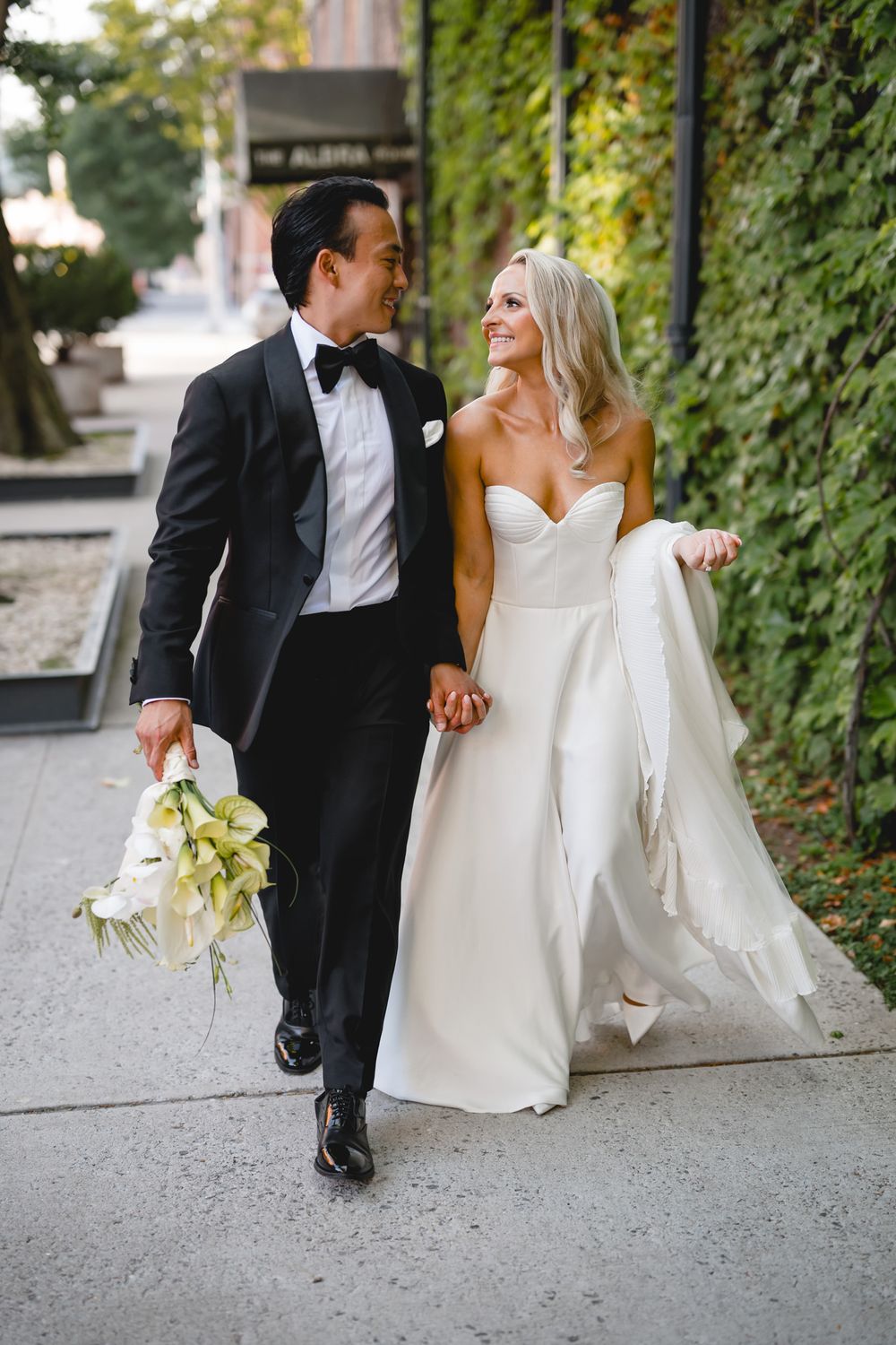 Smiling bride and groom walk hand-in-hand on a city sidewalk, bride holding her gown and groom carrying white calla lilies.