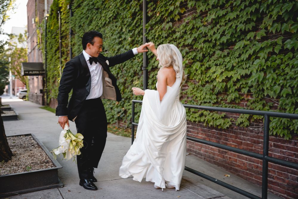 Groom in tuxedo twirling bride in white gown on sidewalk beside ivy-covered brick wall.