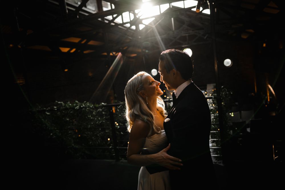 A bride and groom share a romantic moment in dramatic backlit light at an elegant indoor wedding venue.