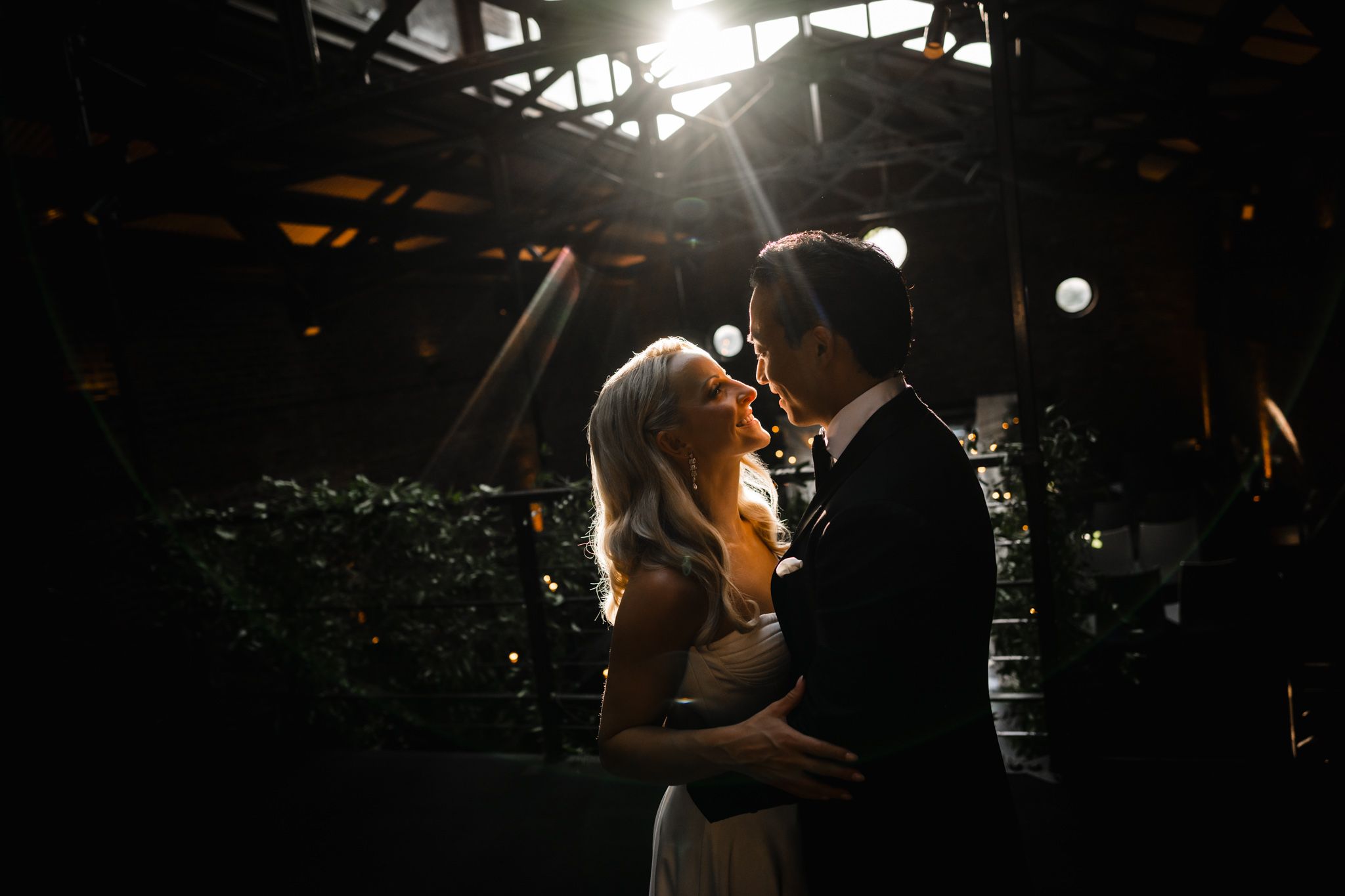 A bride and groom share a romantic moment in dramatic backlit light at an elegant indoor wedding venue.