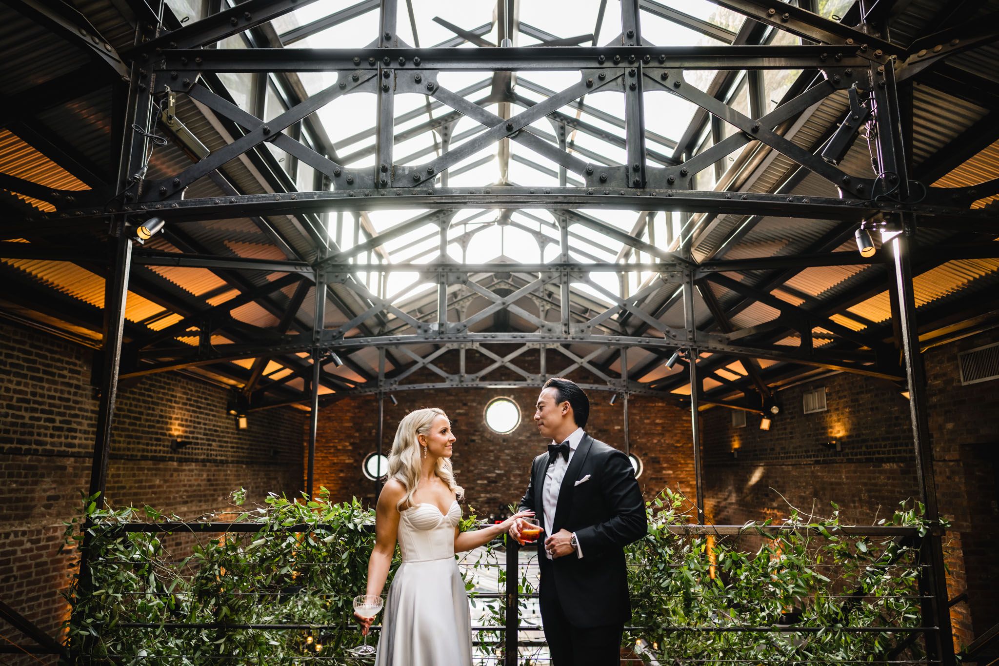 Bride and groom share a joyful moment under industrial glass ceiling with exposed steel beams and greenery.
