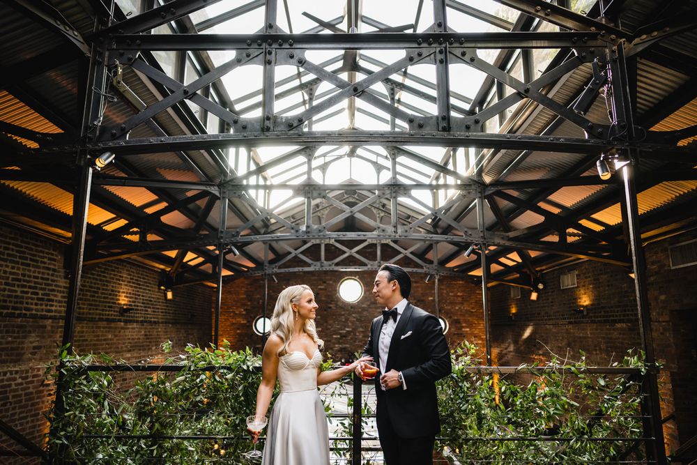 Bride and groom share a joyful moment under industrial glass ceiling with exposed steel beams and greenery.