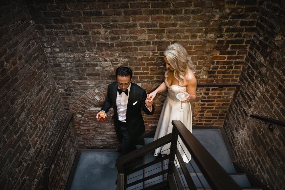 Bride and groom walk down a staircase holding hands in a brick-walled venue.