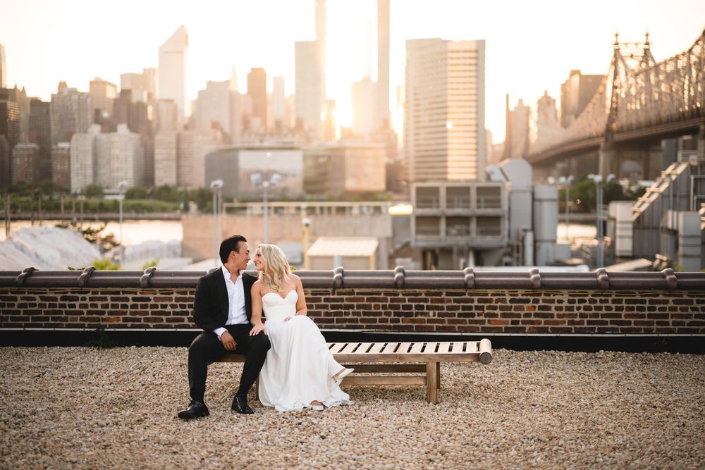 Bride and groom sit on a bench on a rooftop with NYC skyline and golden sunset backdrop.