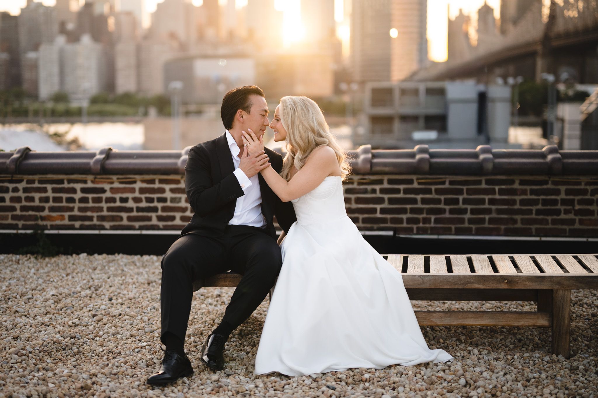 Bride and groom share a romantic kiss on a bench with NYC skyline and golden sunset backdrop.