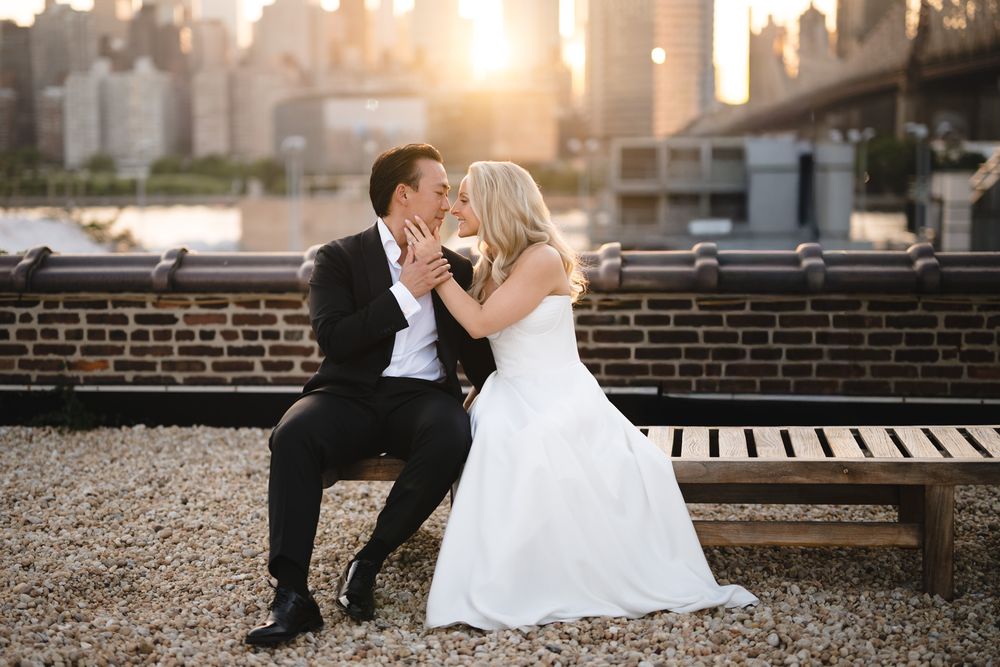 Bride and groom share a romantic kiss on a bench with NYC skyline and golden sunset backdrop.