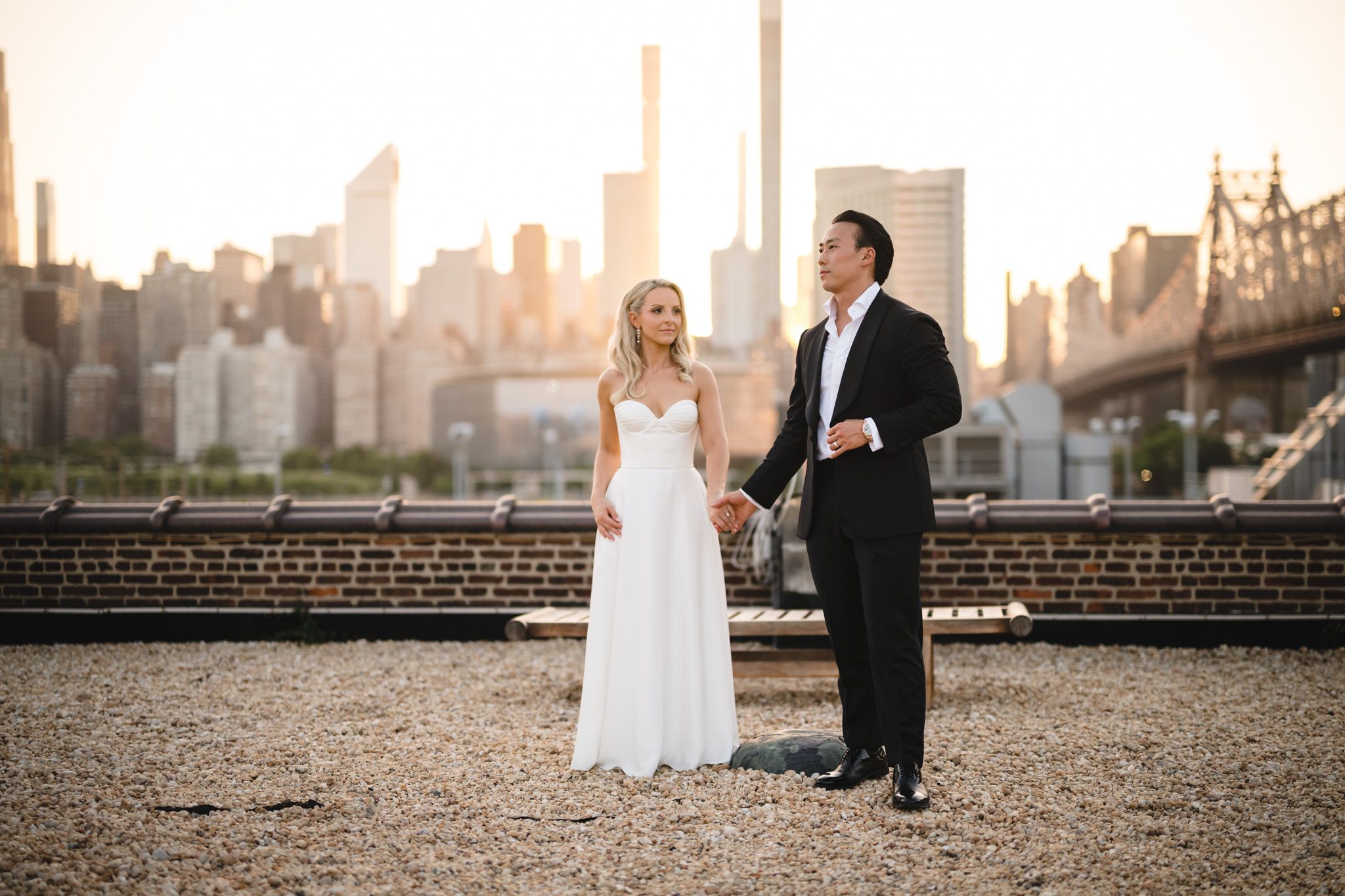 Bride and groom holding hands on rooftop with NYC skyline and Queensboro Bridge at golden hour.