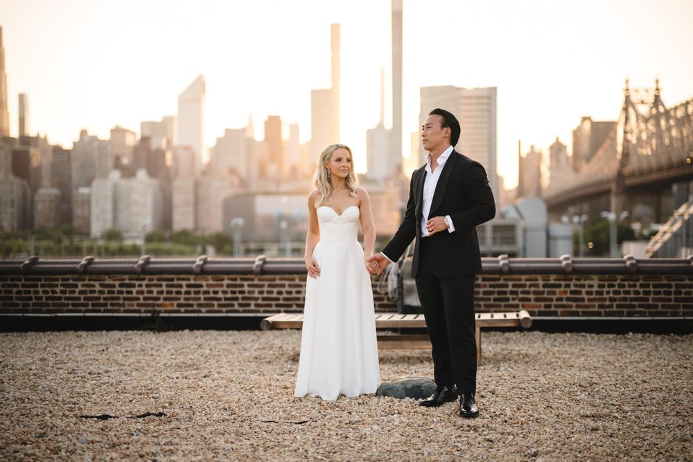 Bride and groom holding hands on rooftop with NYC skyline and Queensboro Bridge at golden hour.