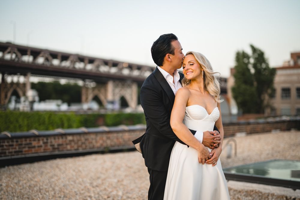 Groom embracing bride from behind, kissing her cheek at golden hour outdoor wedding venue.
