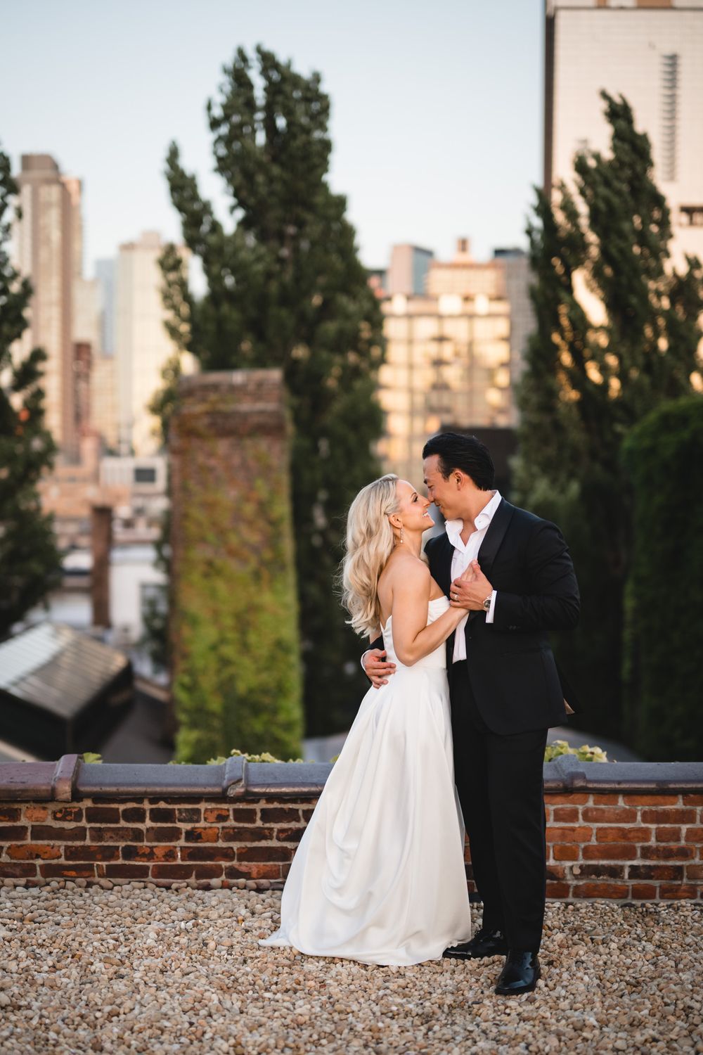 Bride and groom sharing a romantic kiss on rooftop with city skyline and tall trees behind.
