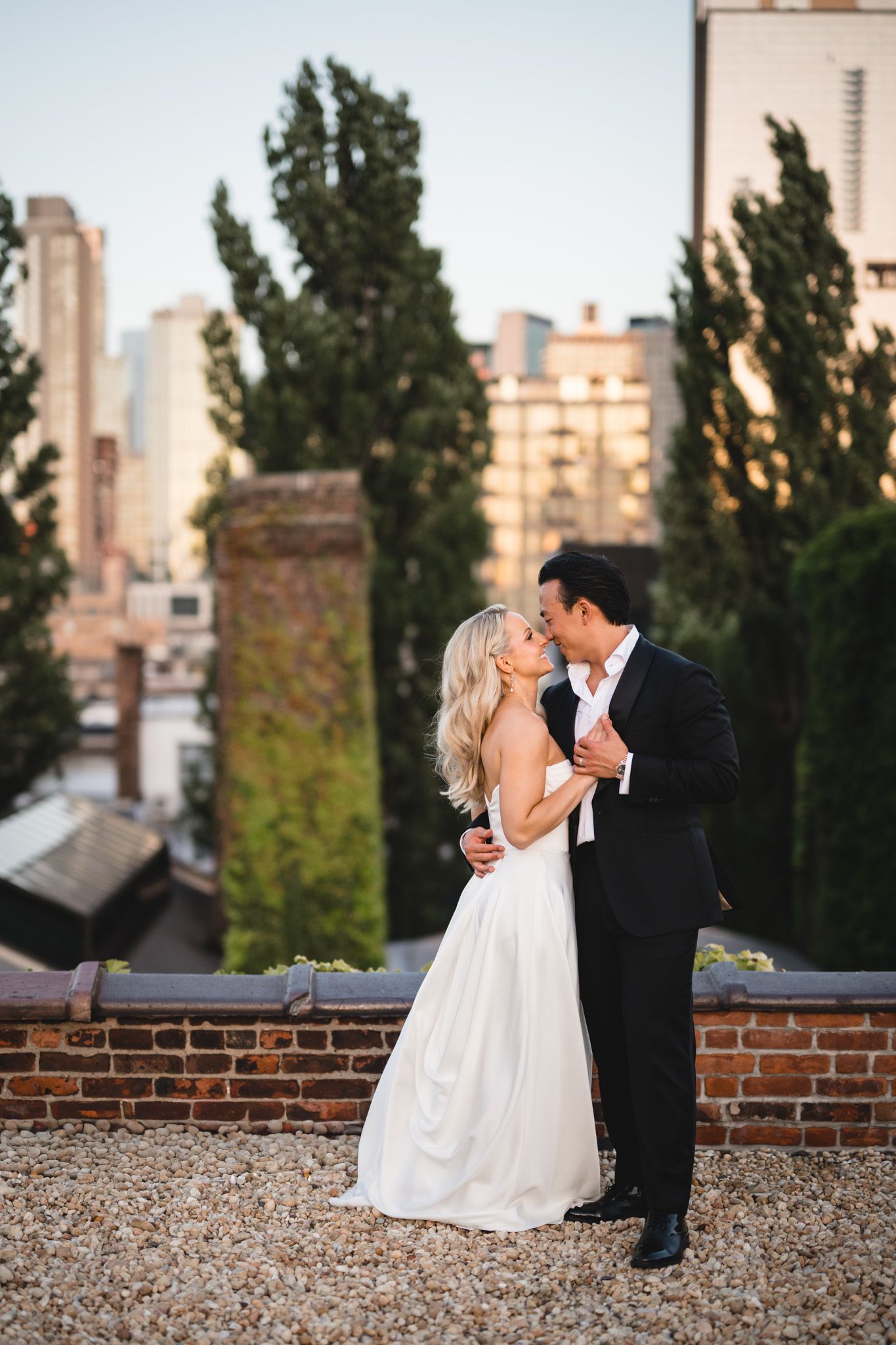 Bride and groom sharing a romantic kiss on rooftop with city skyline and tall trees behind.