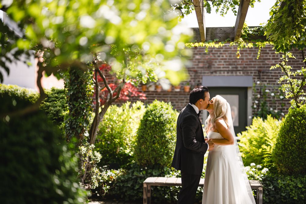 Bride and groom share romantic kiss in lush garden surrounded by green hedges and trees.