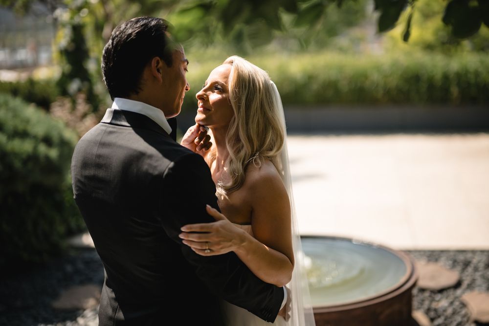 Bride and groom share a romantic embrace outdoors near a fountain, with lush greenery in the background.
