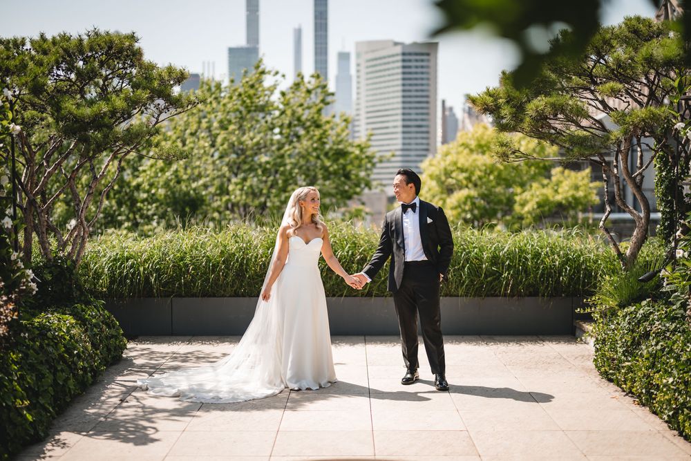 Bride and groom hold hands on rooftop garden with city skyline backdrop on wedding day.
