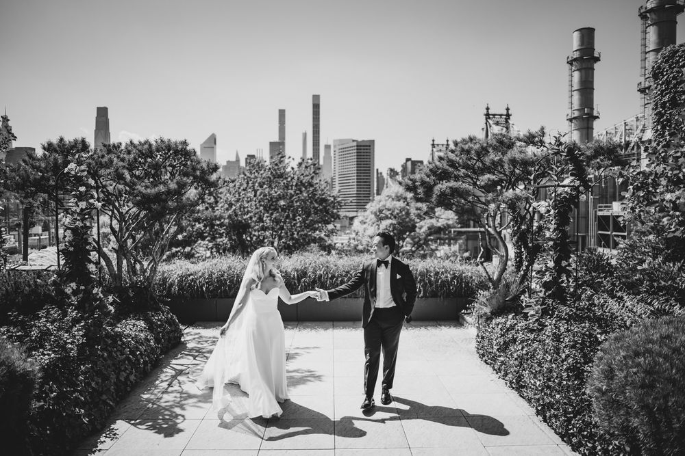 Black and white photo of bride and groom holding hands on a rooftop garden with city skyline backdrop.