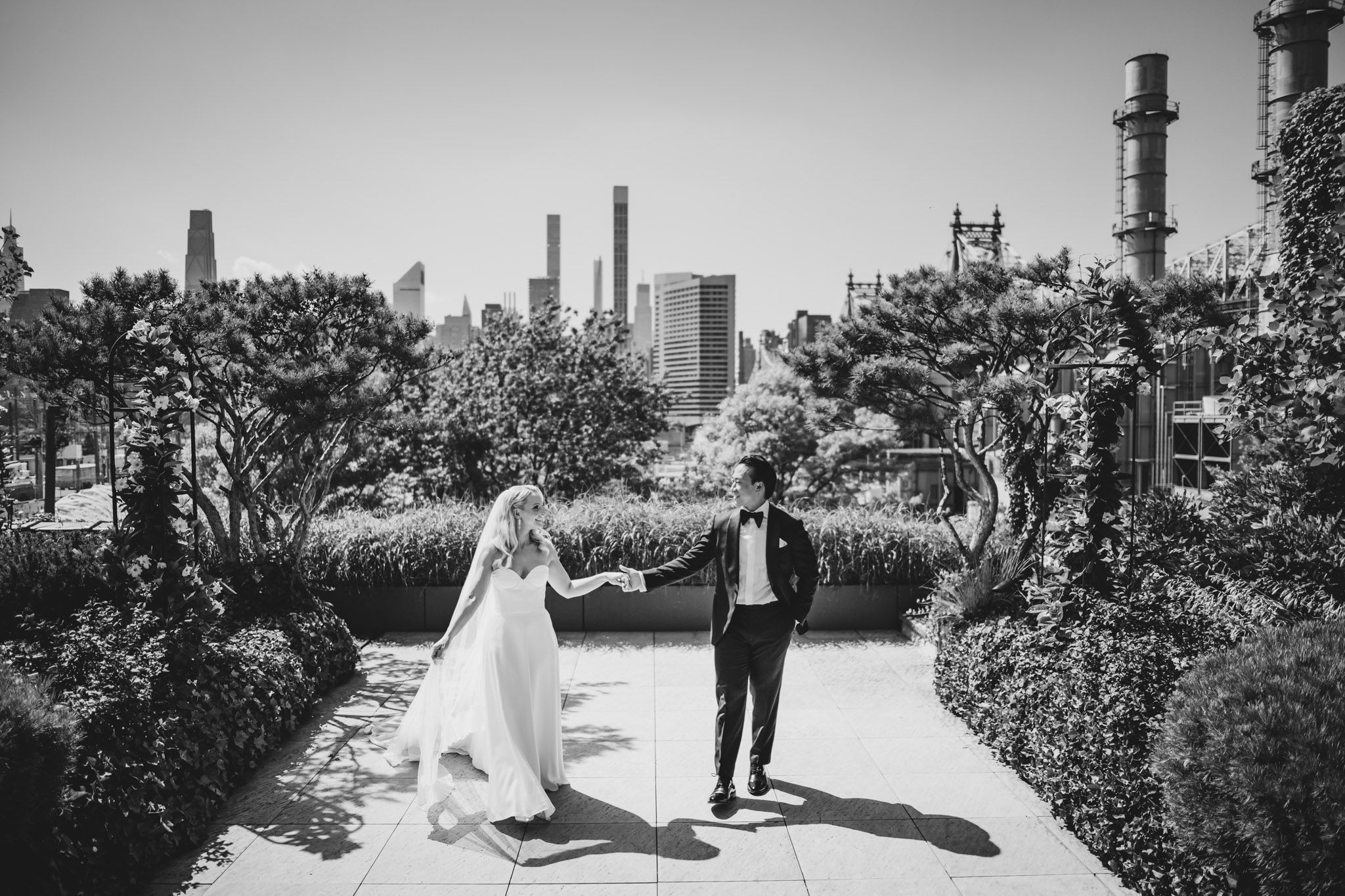Black and white photo of bride and groom holding hands on a rooftop garden with city skyline backdrop.