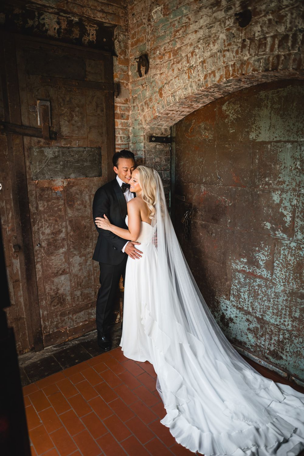 Bride in white gown and veil embraces groom in black tuxedo in rustic brick doorway.