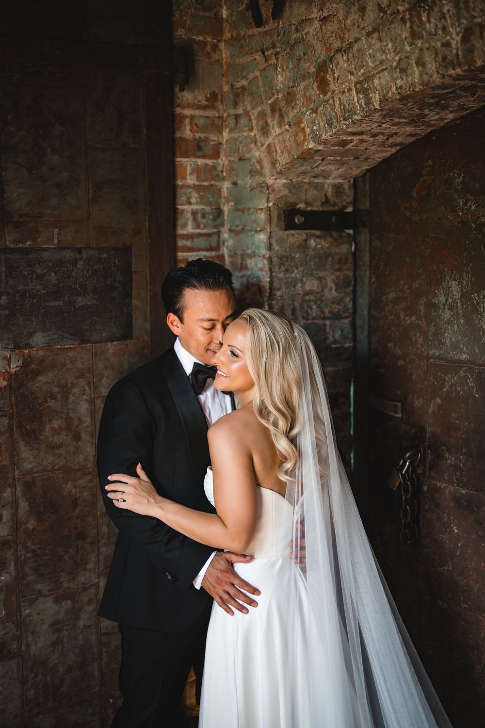 Bride and groom share romantic embrace in rustic brick doorway, bride in strapless white gown with veil, groom in black tuxedo.