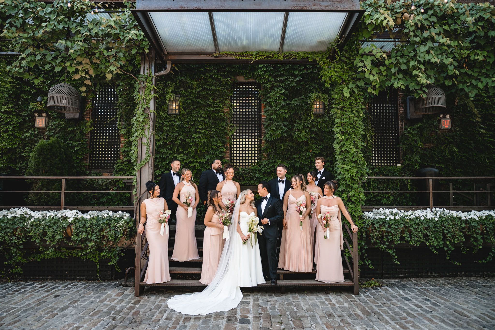 Wedding party poses before an ivy-covered building facade on cobblestone street.