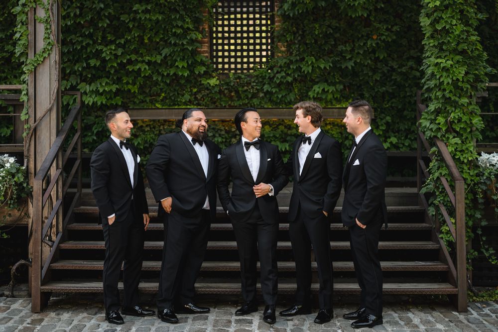 Five groomsmen in black tuxedos with bow ties posing on stone steps surrounded by lush ivy walls.
