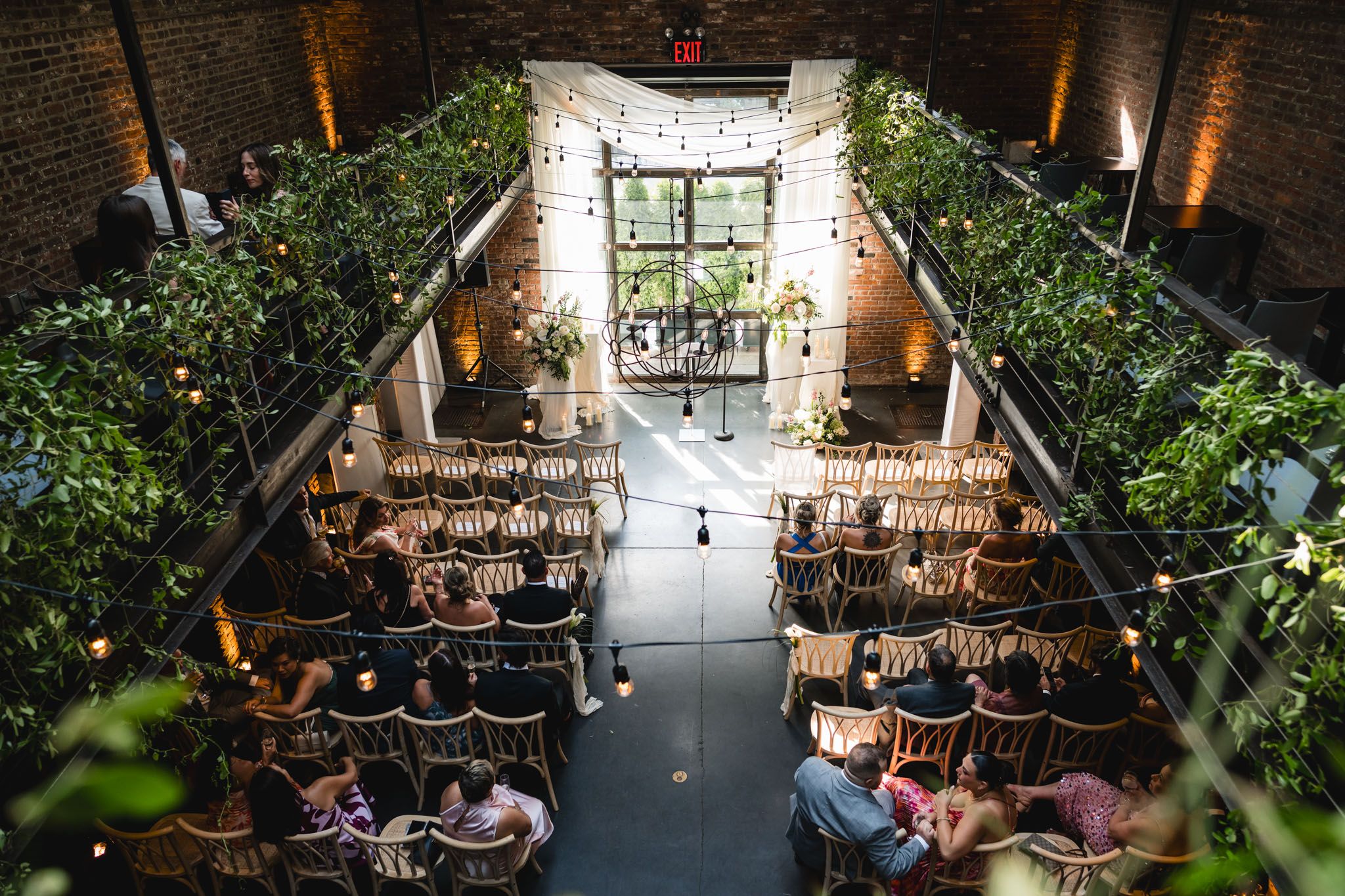 Aerial view of elegant wedding ceremony in brick venue with string lights and lush greenery.