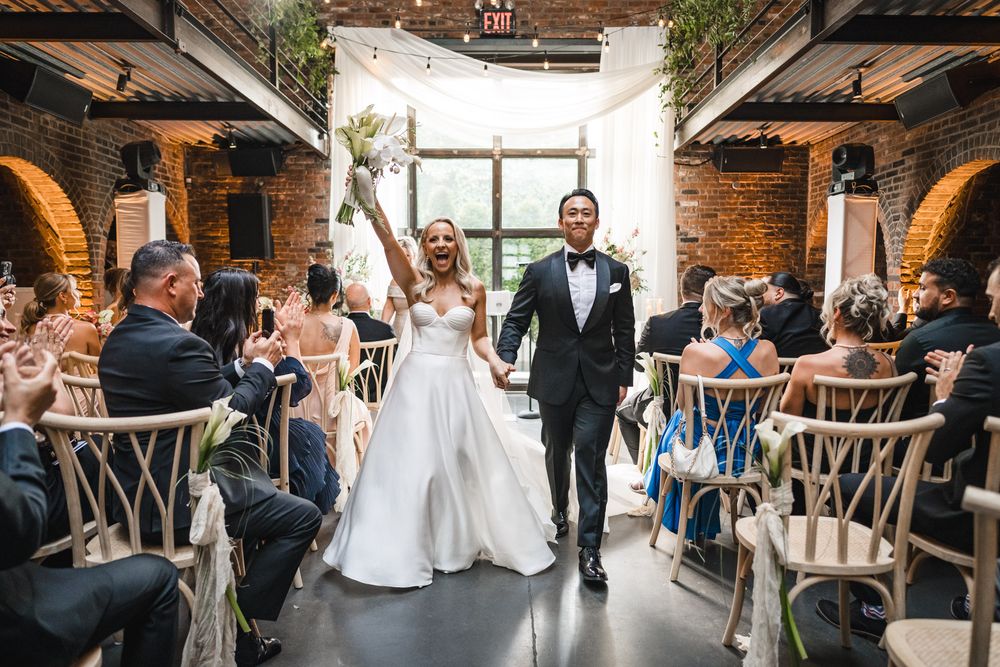 Joyful newlyweds celebrate recessional in rustic brick venue, bride raising bouquet, guests applauding in elegant wedding ceremony.