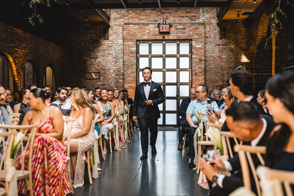 Groom walking down the aisle in elegant brick-walled wedding venue with seated guests watching.