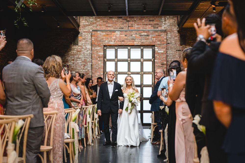 Bride walks down the aisle with her father in a rustic brick-walled wedding venue with guests watching.