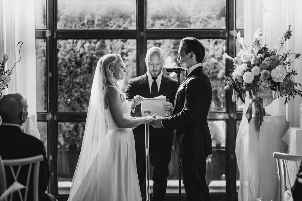 Black and white wedding ceremony photo of bride and groom exchanging vows before officiant near large windows.