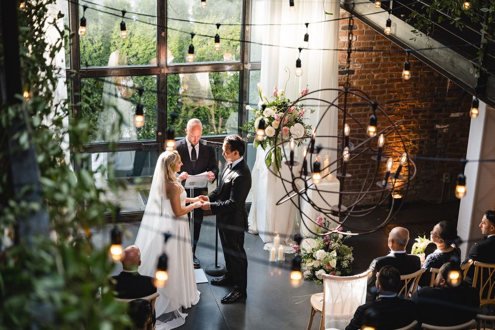 Couple exchanging vows at an elegant indoor wedding ceremony with brick walls, string lights, and floral arch backdrop.