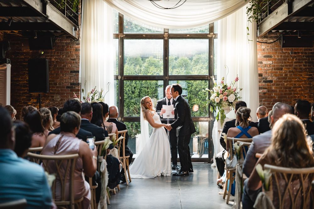 Bride and groom share first kiss at indoor wedding ceremony with brick walls and large windows overlooking lush greenery.