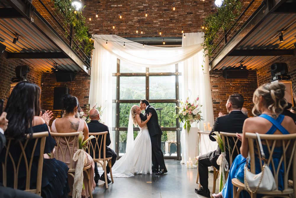 Bride and groom share first kiss at elegant indoor wedding ceremony with string lights and floral arch.