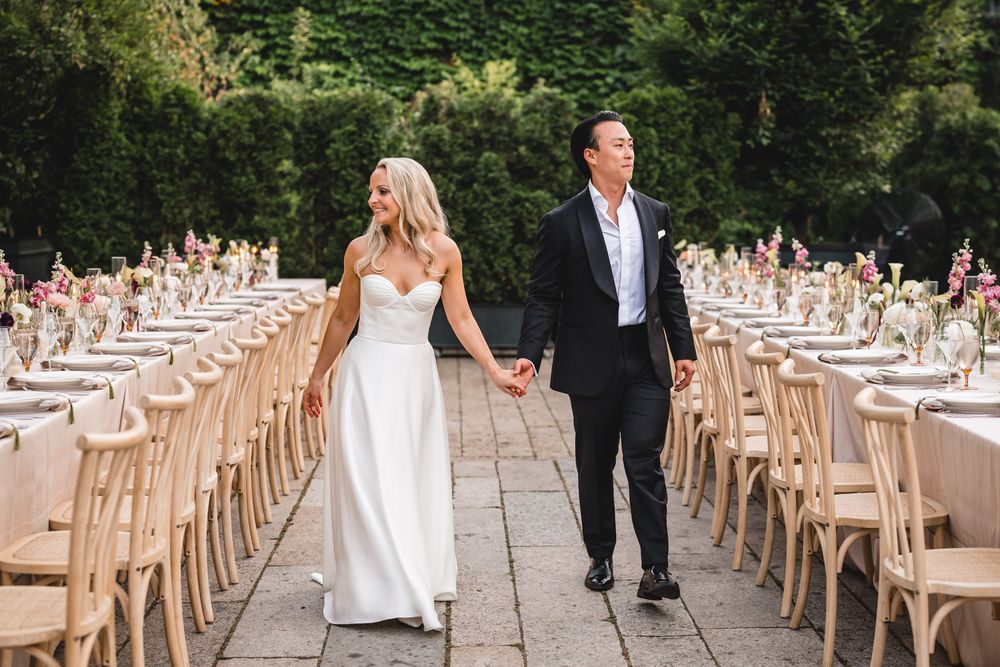 Bride and groom walk hand-in-hand between elegantly set long tables at an outdoor wedding reception.