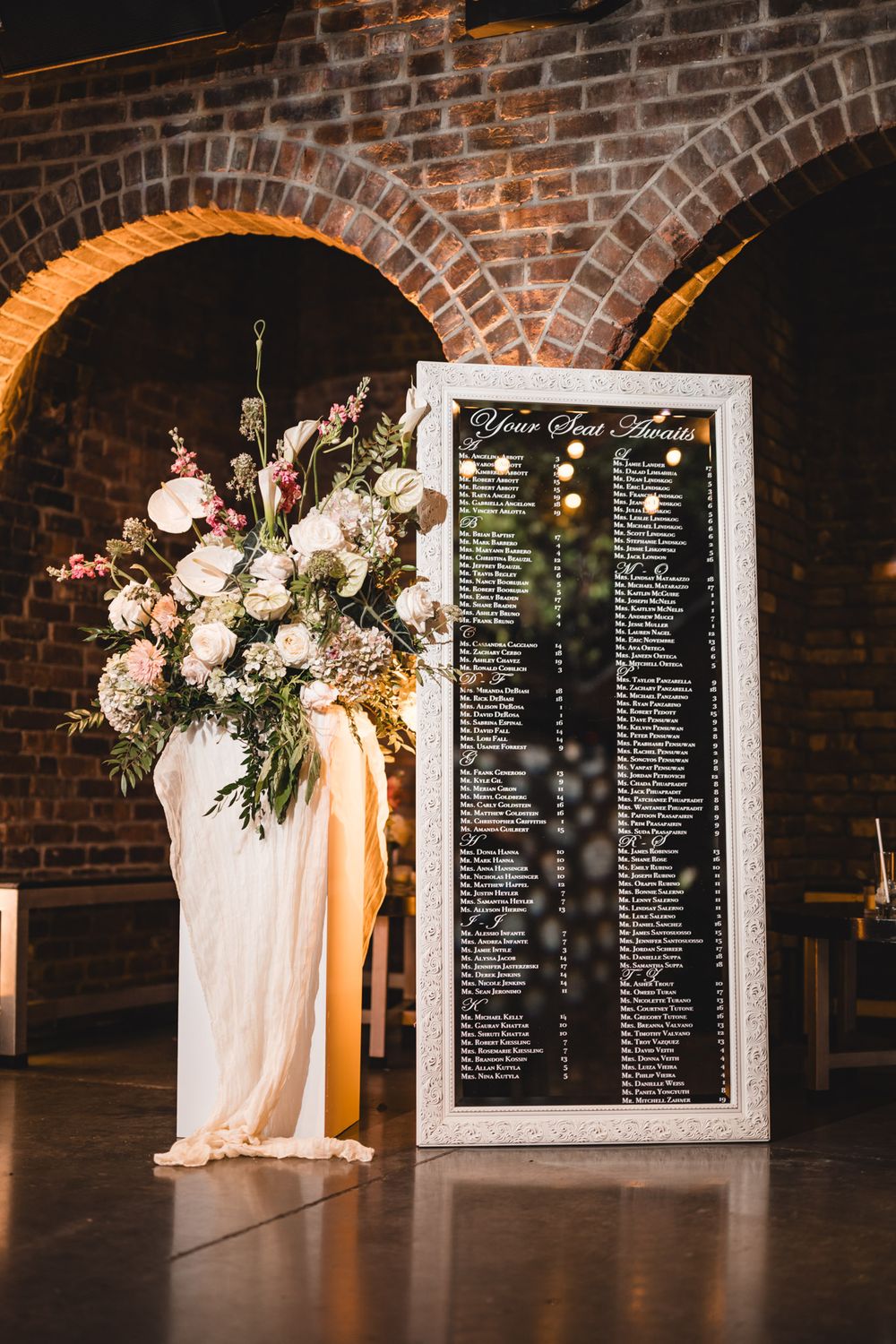 Wedding seating chart in ornate white frame beside large floral arrangement in rustic brick arch venue.
