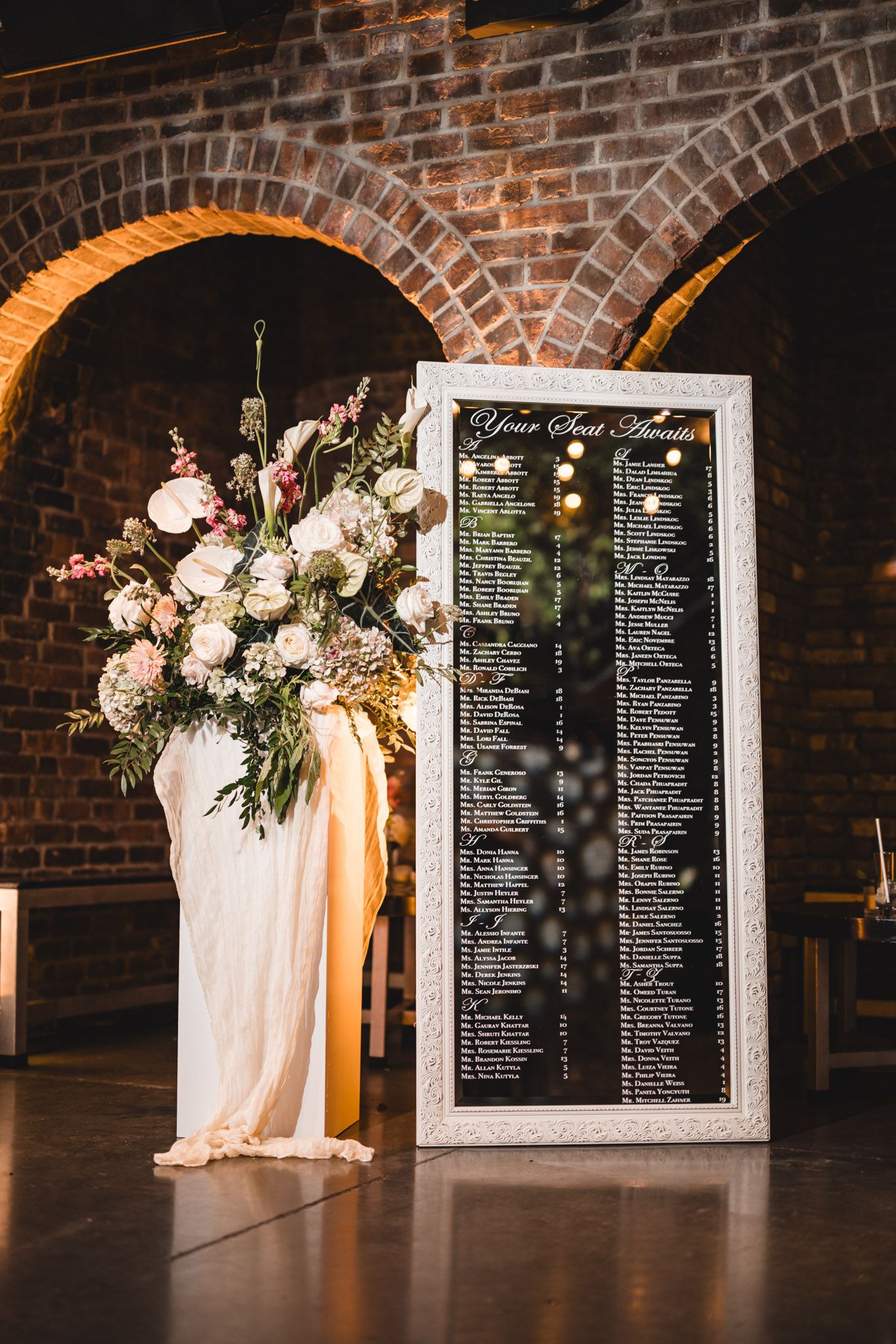 Wedding seating chart in ornate white frame beside large floral arrangement in rustic brick arch venue.