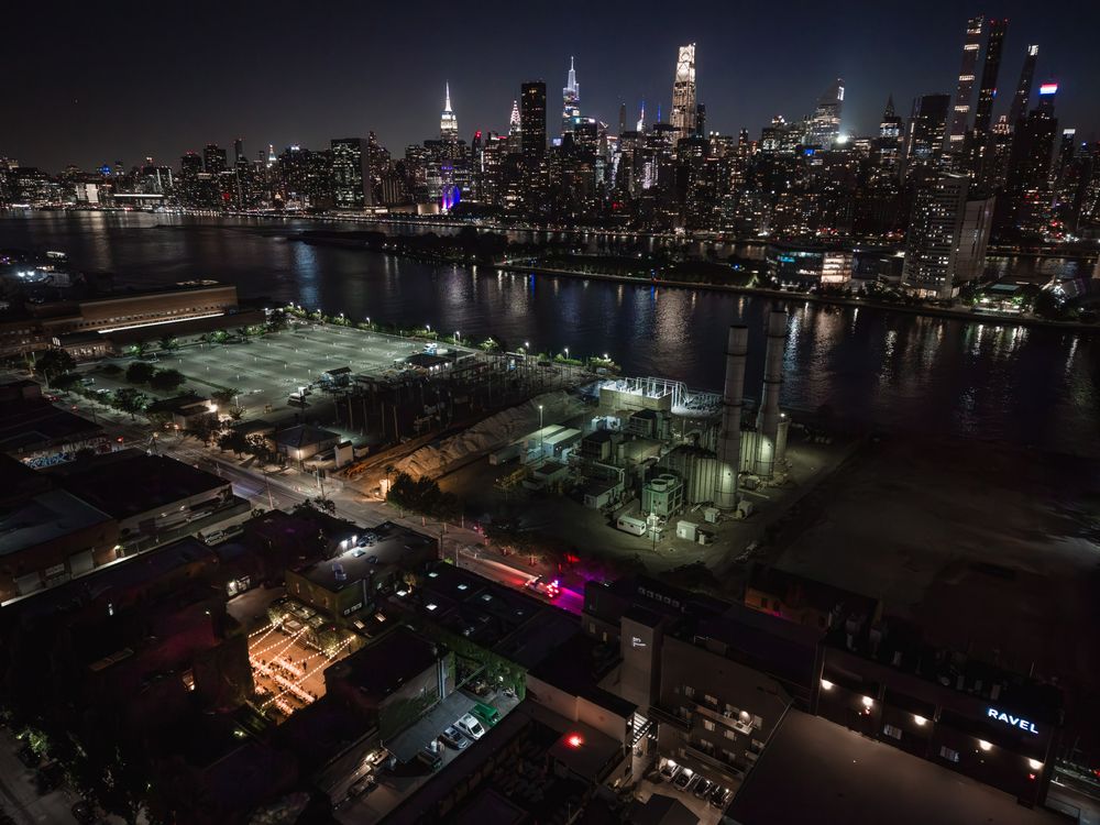 Aerial night view of NYC skyline across the East River with industrial waterfront and glowing city lights.