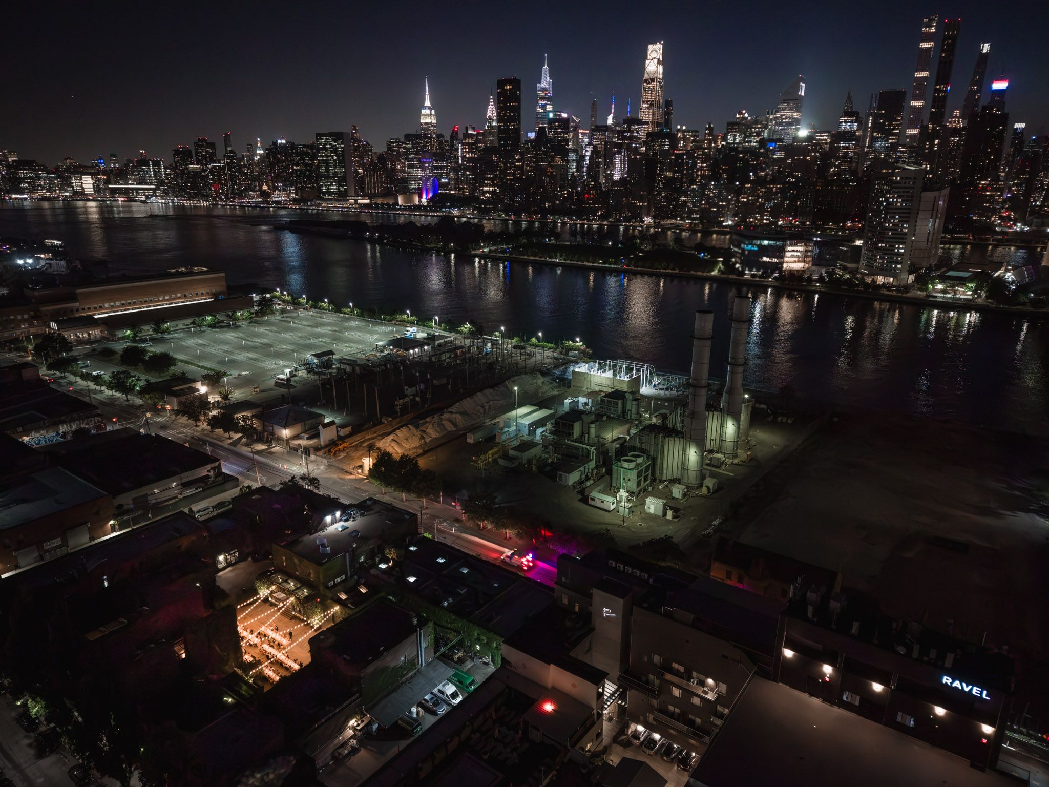 Aerial night view of NYC skyline across the East River with industrial waterfront and glowing city lights.