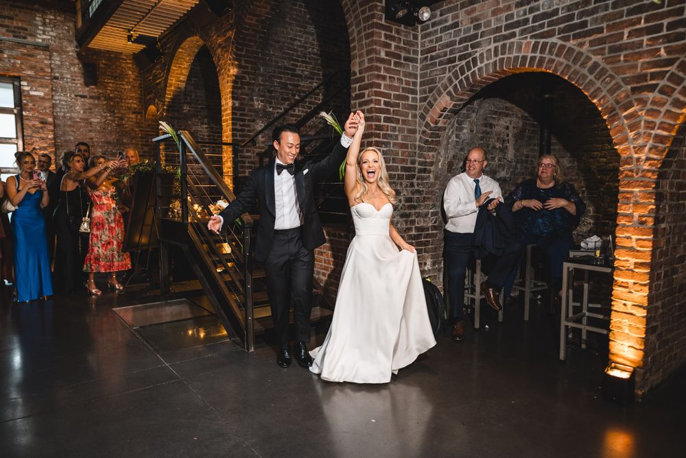 Bride and groom make grand entrance at rustic brick venue, raising hands in celebration as guests cheer.