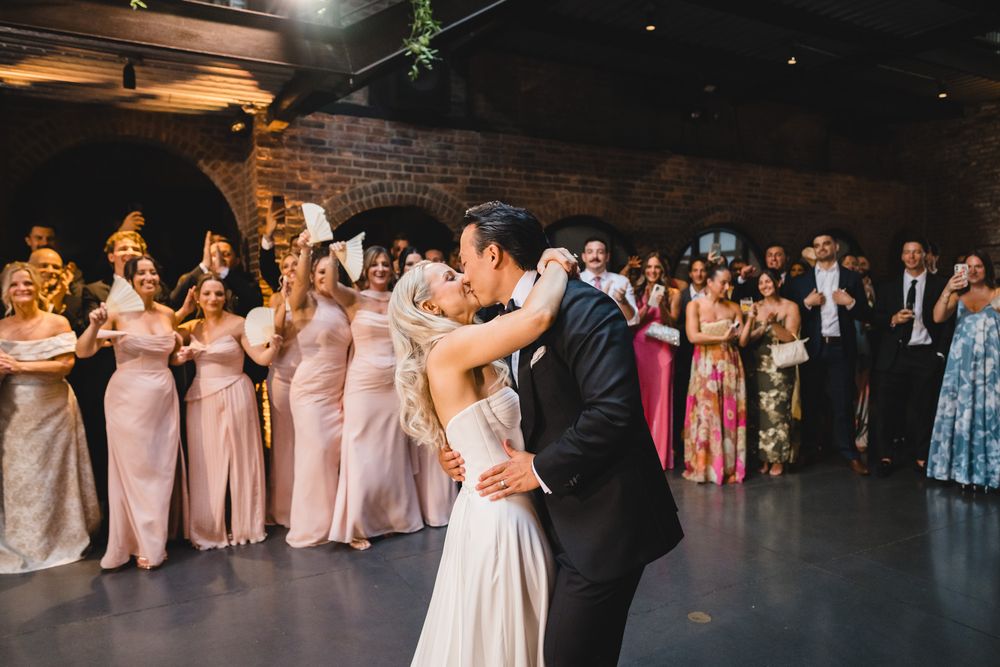 Bride and groom share first dance kiss surrounded by bridesmaids in pink gowns at indoor wedding reception.
