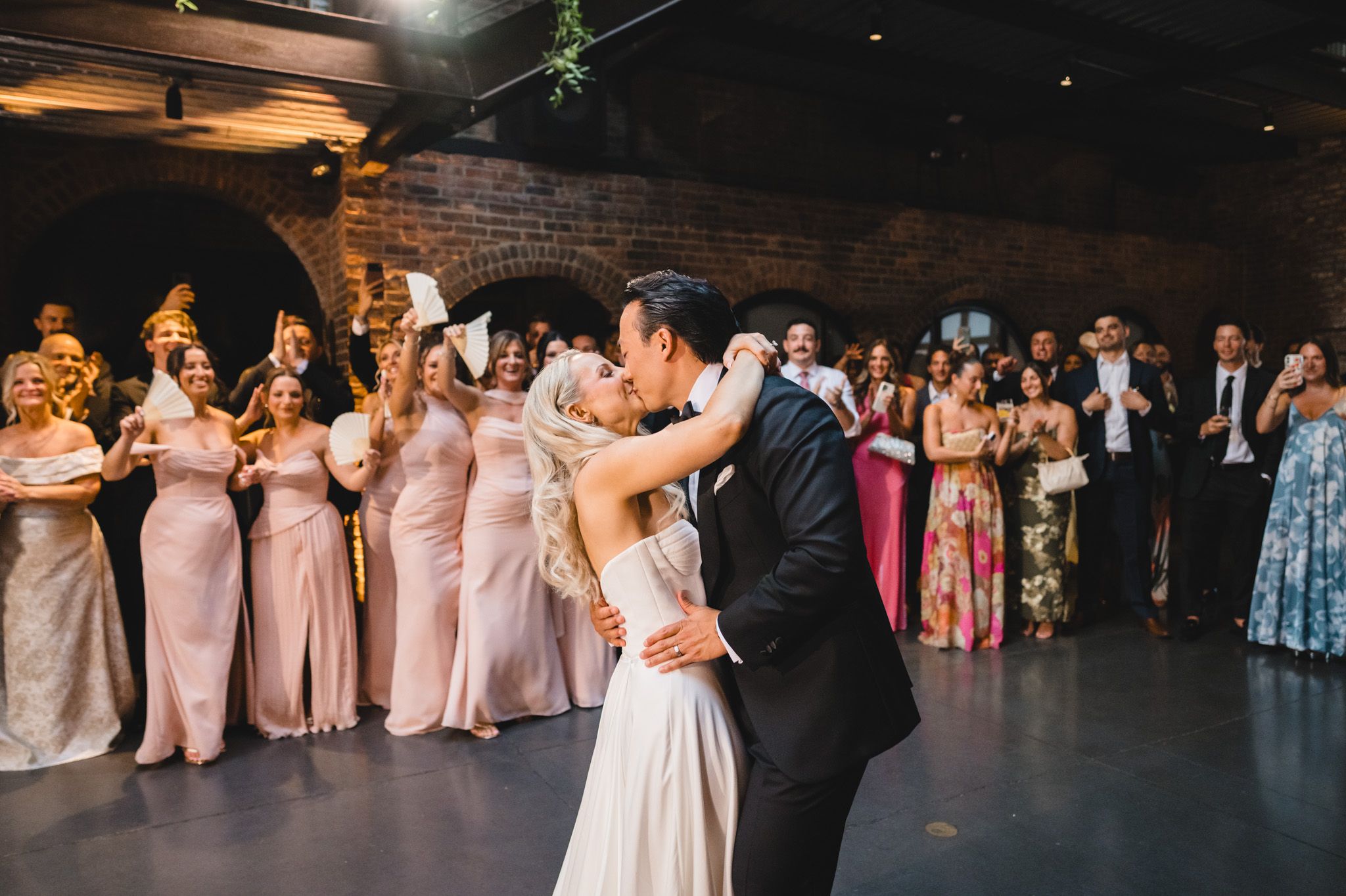 Bride and groom share first dance kiss surrounded by bridesmaids in pink gowns at indoor wedding reception.